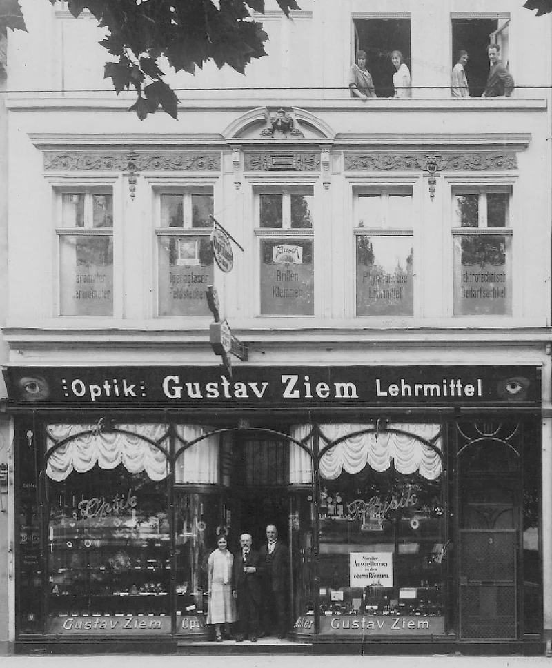 "Historisches Foto des Optikgeschäfts Gustav Ziem. Die Fassade zeigt dekorative Verzierungen und Schaufenster mit Brillen und Lehrmitteln. Drei Personen stehen vor dem Eingang, und im oberen Stockwerk schauen Menschen aus geöffneten Fenstern."